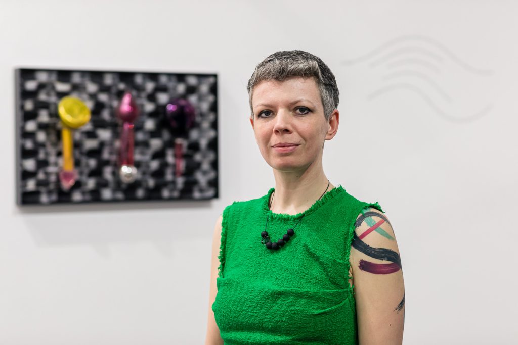 Photo of Vicky from the shoulders up, behind her is a display of her glass sculptures on the wall