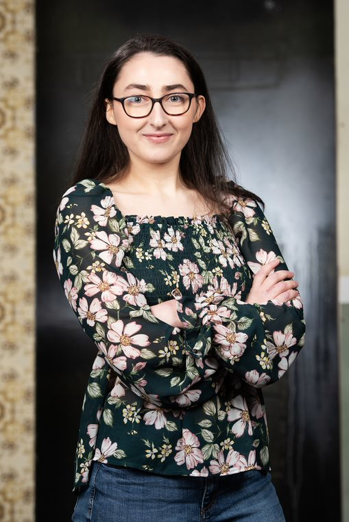 A photo of Joanna stood in a doorway with her arms folded, smiling at the camera. She is wearing a black top with a flower pattern and wears glasses.
